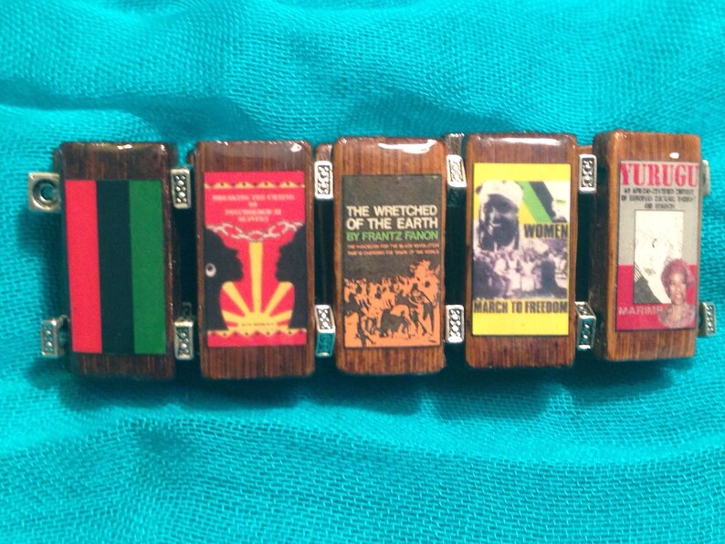 May include: A wooden bracelet with six charms, each featuring a different image. The charms are decorated with red, green, black, yellow, and white colours. The images include a flag, a book cover, a group of people, a woman's face, and a book cover with the word "YURUGU".