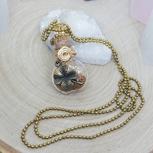 Puede incluir: Un collar de cadena dorada con un pequeño colgante de botella de vidrio lleno de purpurina dorada y un amuleto de trébol de cuatro hojas.