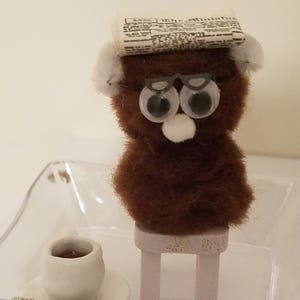 May include: A brown fuzzy toy bear wearing glasses and a newspaper on its head sits on a small white stool. The bear is holding a small white cup with a red liquid inside. The bear and cup are sitting on a clear plastic tray.