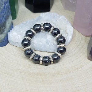 Puede incluir: Un brazalete de cuentas de hematites negras con detalles plateados. El brazalete está hecho de cuentas redondas y está sobre una superficie blanca.