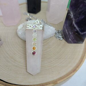 Könnte beinhalten: Ein Anhänger aus rosa Rosenquarz mit einer silbernen Fassung und sieben Chakra-Steinen. Die Steine sind in einer Linie in der Mitte des Anhängers angeordnet. Der Anhänger hängt an einer silbernen Kette.