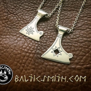May include: Two silver axe pendants with intricate star-shaped designs on a brown leather background. The pendants are on chains and have a hole at the top for hanging. The text "BALTICSMITH.COM" is visible at the bottom of the image.
