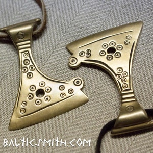 May include: Two bronze-colored pendants in the shape of axes, each with a series of small, circular indentations. The pendants are connected to brown and black cords. The text "BALTICSMITH.COM" is visible.