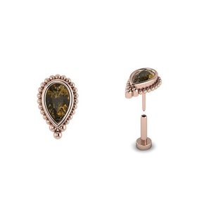 May include: A teardrop-shaped body jewellery piece with a clear, faceted gemstone set in a rose gold bezel. The gemstone is surrounded by a beaded halo. The jewellery piece is attached to a rose gold post with a threaded end.