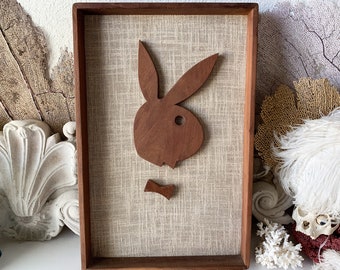 May include: A framed wall art piece featuring a brown wooden cutout of the Playboy bunny logo on a beige textured background.
