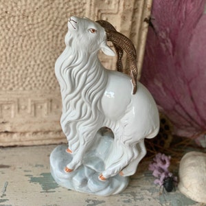 May include: White porcelain figurine of a goat with brown horns standing on a gray base. The goat is looking up and has a detailed, textured fur.