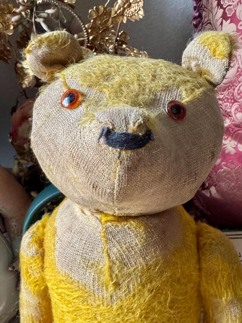 May include: A vintage teddy bear with a tan head and yellow fur. The bear has orange eyes and a black stitched nose. The bear's fur is worn, giving it a classic, antique appearance. The bear's head is made of a different material than the body.