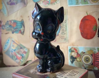 May include: A black ceramic cat figurine with large eyes and a red nose. The cat is sitting on a book with the word "Triumph" visible.