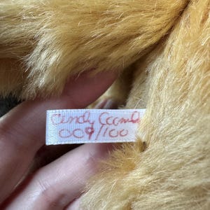 May include: Close-up of a plush toy with golden-brown fur. A white tag with red text reads "Cindy Ceech" and "009/100". The fur is soft and fluffy, with a slight sheen. The toy appears to be well-made and in good condition.