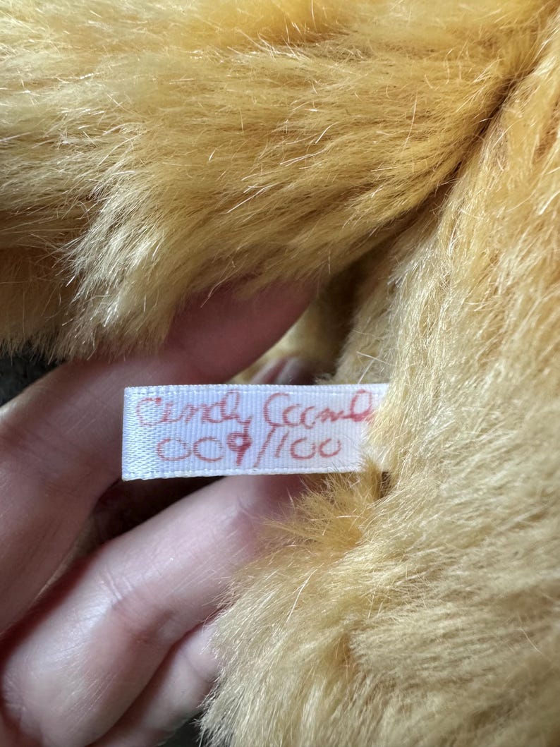 May include: Close-up of a plush toy with golden-brown fur. A white tag with red text reads "Cindy Ceech" and "009/100". The fur is soft and fluffy, with a slight sheen. The toy appears to be well-made and in good condition.