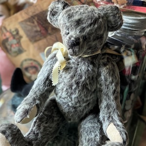 May include: A vintage-style teddy bear with gray fur and a cream-colored bow around its neck. The bear has a classic design with a slightly worn appearance, giving it a nostalgic feel. The bear is sitting, with its paws and ears visible.