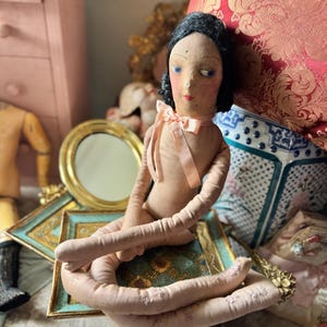 May include: A vintage-style cloth doll with black hair, blue eyes, and a pink bow. The doll is seated on a decorative frame with a gold-rimmed mirror. Other antique items are in the background, including a wooden mannequin and a pink dresser.