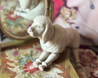 May include: A vintage-style, off-white poodle figurine with a textured coat and pink accents on the nose and mouth. The dog is posed in a seated position. The figurine is on a floral patterned surface.
