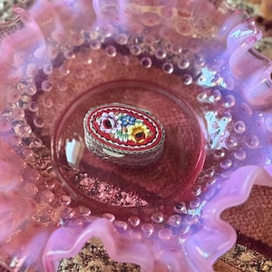 May include: An oval, silver-toned trinket box with a mosaic floral design in red, yellow, blue, and white. The box sits inside a pink, ruffled glass dish. The dish has a clear, textured rim and a smooth, reflective surface.