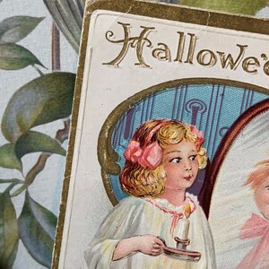 Pode incluir: Postal vintage de Halloween com uma menina em um vestido branco segurando uma vela. O cart&atilde;o tem detalhes dourados e a palavra "Halloween" no topo. O fundo inclui um design floral.