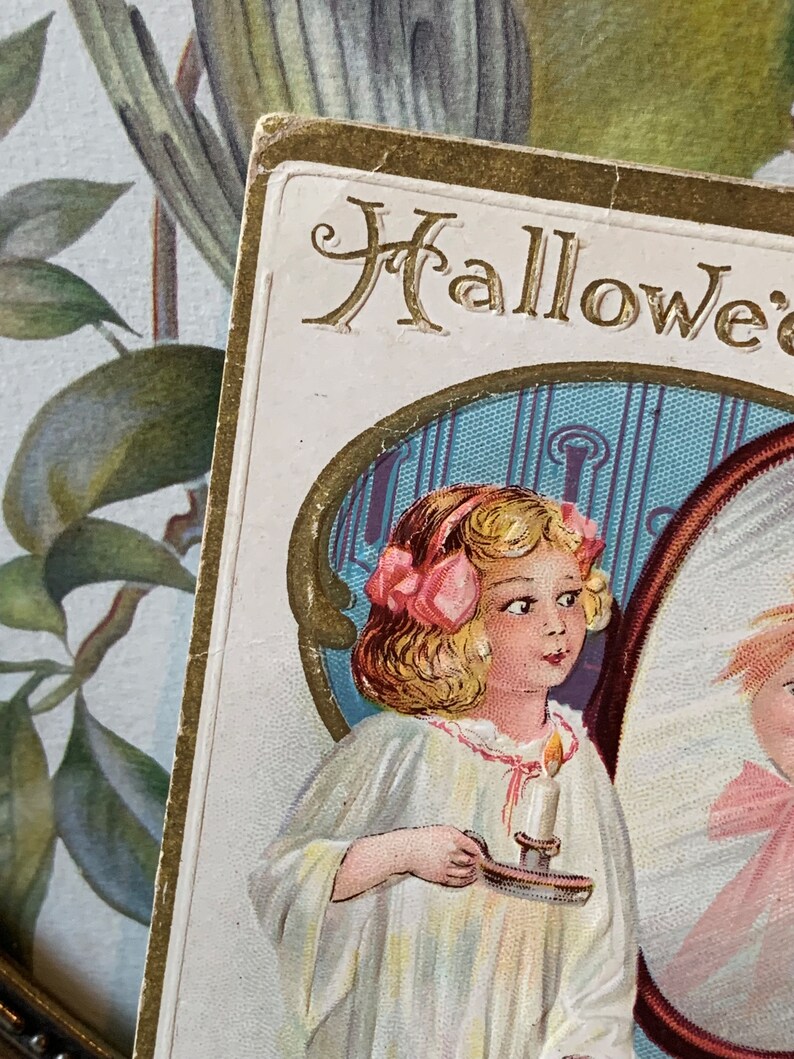 Pode incluir: Postal vintage de Halloween com uma menina em um vestido branco segurando uma vela. O cart&atilde;o tem detalhes dourados e a palavra "Halloween" no topo. O fundo inclui um design floral.