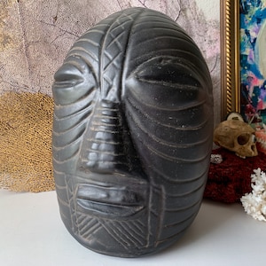 May include: A black ceramic sculpture of a face with detailed carvings. The face has a closed eye, a prominent nose, and a carved mouth.
