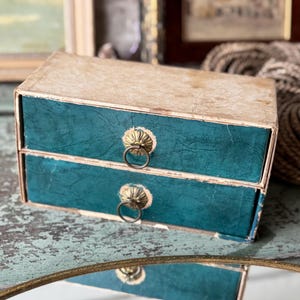 May include: A small, vintage two-drawer box with a cream-colored top and teal-colored drawers. The drawers have ornate brass pulls with ring handles. The box is likely used for storing small items.