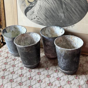 May include: A set of four cylindrical metal cups featuring a weathered, dark gray finish with visible white distressed textures on the exterior and interior surfaces. Each cup has a simple, open-top design with a slightly flared rim and a rounded base.
