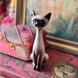 May include: A ceramic figurine of a Siamese cat with blue eyes. The cat is sitting with its tail curled around its legs. The figurine is painted in shades of brown and white.