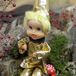 May include: A vintage doll with blonde hair, blue eyes, and an open mouth, wearing a gold outfit with a black collar and a matching hat. The doll is posed in a natural setting with rocks, moss, and red berries.