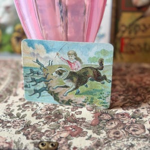 May include: A rectangular vintage card features a child with a dog chasing black cats. The illustration has a blue sky and green grass. The card is set against a floral patterned fabric and a pink glass object.