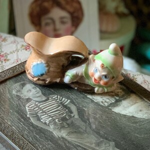 May include: A vintage ceramic figurine of a whimsical elf with a green hat and blue eyes, next to a small brown pitcher with a blue patch. The figurine is resting on a framed black and white photograph.