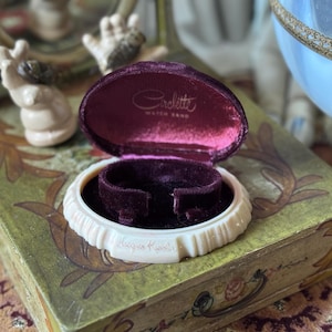 May include: An open, vintage watch band box with a cream-colored, scalloped exterior and a deep burgundy velvet interior. The box is labeled "Circlette Watch Band" and sits on a decorative, green and gold painted box.