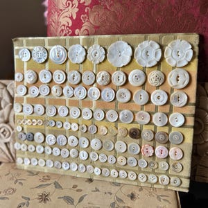 May include: A collection of vintage buttons arranged on a rectangular display board. The buttons vary in size, shape, and color, including white, pearl, and brown tones. The board has a yellow and gold fabric background.