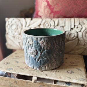May include: A cylindrical ceramic planter with a textured, bark-like exterior and embossed floral designs. The planter has a green interior and is painted in shades of blue and gray. The planter is sitting on a floral patterned box.