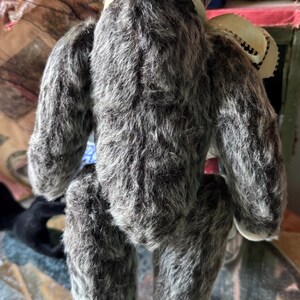 May include: A handmade gray teddy bear with a cream-colored ribbon around its neck. The bear has a fluffy, textured appearance and is posed from the back. The bear's fur is a mix of gray and white, giving it a soft, vintage look.