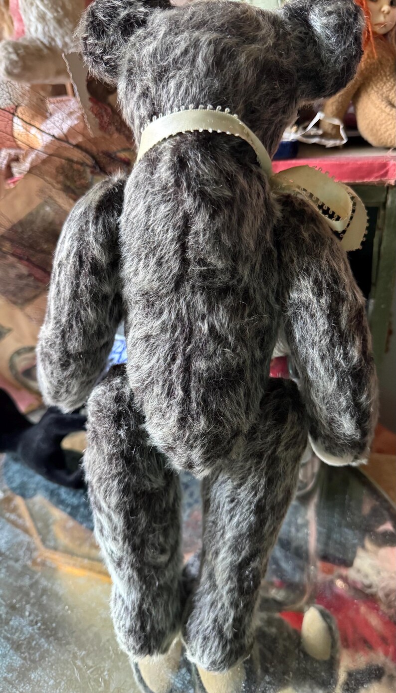 May include: A handmade gray teddy bear with a cream-colored ribbon around its neck. The bear has a fluffy, textured appearance and is posed from the back. The bear's fur is a mix of gray and white, giving it a soft, vintage look.