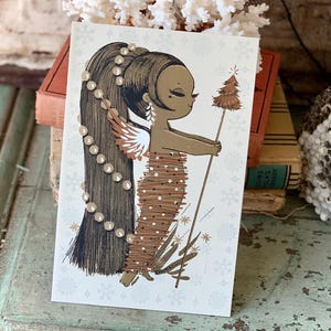 May include: A vintage Christmas card featuring a brown-skinned angel with long black hair adorned with white beads. The angel is wearing a brown dress with white polka dots and holding a small, gold-trimmed Christmas tree. The background is white with a snowflake pattern.