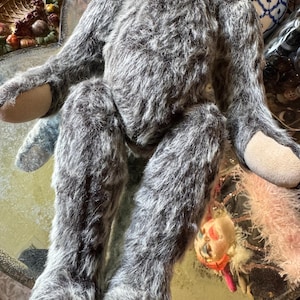 May include: A handmade, gray plush toy with long limbs and a furry texture. The toy has tan-colored pads on its hands and feet. A small, decorative ornament hangs near the toy's legs. The toy is positioned on a reflective surface.