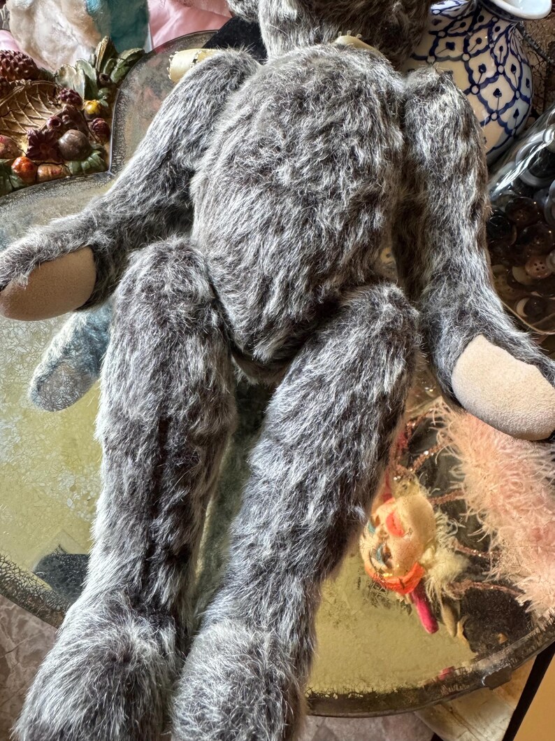 May include: A handmade, gray plush toy with long limbs and a furry texture. The toy has tan-colored pads on its hands and feet. A small, decorative ornament hangs near the toy's legs. The toy is positioned on a reflective surface.