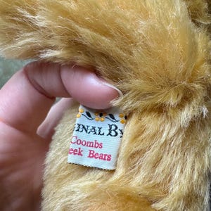 May include: Close-up of a plush, golden-brown teddy bear. The fur is long and textured. A tag reads "Original By Coombs Cheek Bears" in a decorative font. The tag has a white background with a floral design.