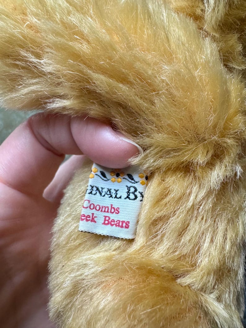 May include: Close-up of a plush, golden-brown teddy bear. The fur is long and textured. A tag reads "Original By Coombs Cheek Bears" in a decorative font. The tag has a white background with a floral design.