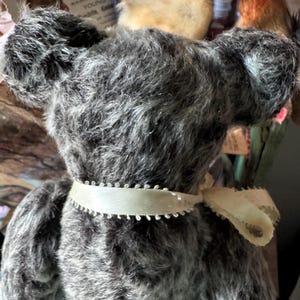 May include: A vintage gray teddy bear with a cream-colored ribbon around its neck. The bear has a fluffy, textured appearance and large, rounded ears. The ribbon is tied in a bow at the back. The bear is part of a collection of antique toys.