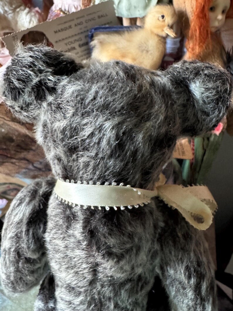 May include: A vintage gray teddy bear with a cream-colored ribbon around its neck. The bear has a fluffy, textured appearance and large, rounded ears. The ribbon is tied in a bow at the back. The bear is part of a collection of antique toys.