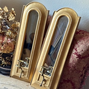 May include: Two gold-colored wall sconces with arched mirrors and candle holders. The sconces have a rectangular shape with an arched top and a decorative border. Each sconce has a small candle holder and a decorative hook.