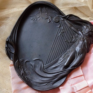 May include: A black metal tray with a raised design of a woman holding a harp. The tray is oval shaped and has a smooth surface.