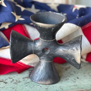 May include: A black, handmade, cross-shaped ceramic sculpture with a flared top and base. The sculpture has a hole in the center and is accented with thin, light-colored lines. The background features an American flag.