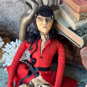 May include: A vintage doll with a black velvet hat, red jacket, and black ribbon. The doll has a painted face with blue eyes and red lips. The doll is sitting on a wooden surface with books stacked behind it.