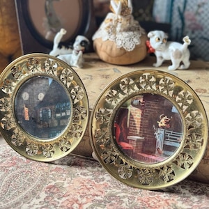 May include: Two circular brass frames featuring intricate openwork leaf patterns along the edges. Each frame holds a small, circular photograph depicting a scene of a building exterior and a brick fireplace interior, respectively.