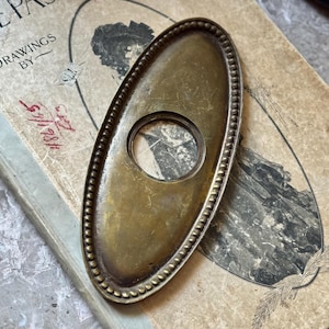 May include: An oval, antique brass door plate with a central hole and a beaded edge. The plate rests on a vintage book with the title "The Past" and the text "Drawings By". The book has a faded illustration and handwritten markings.