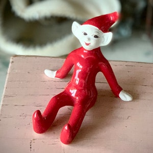 May include: A red ceramic elf figurine with white painted details, sitting with one leg extended and the other bent at the knee.