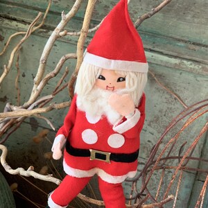 May include: A vintage Christmas Santa Claus doll. The doll is dressed in a red felt suit with white polka dots, a black belt, and white trim. It has a white beard, a red hat, and a smiling face. The doll is a festive holiday decoration.