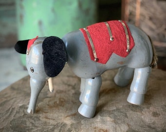 May include: A vintage wooden toy elephant with a gray painted body, black felt ears, and a red felt saddle with silver accents. The elephant has a white painted trunk and tail.