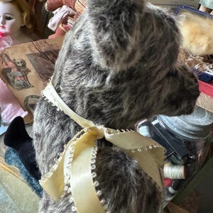 May include: A vintage gray teddy bear with a cream-colored ribbon tied in a bow around its neck. The bear has a soft, plush texture and is surrounded by antique dolls and ephemera, suggesting a collectible or nostalgic theme.