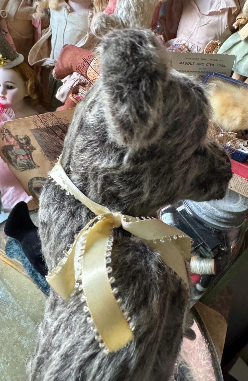 May include: A vintage gray teddy bear with a cream-colored ribbon tied in a bow around its neck. The bear has a soft, plush texture and is surrounded by antique dolls and ephemera, suggesting a collectible or nostalgic theme.